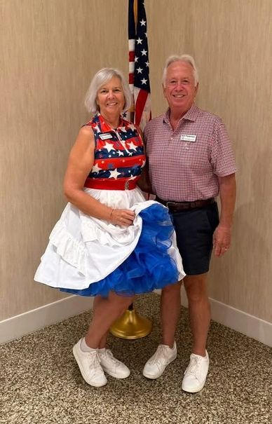 A couple dressed in patriotic red, white, and blue outfits stands smiling by an American flag.