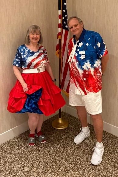 Couple dressed in patriotic red, white, and blue outfits standing by an American flag.