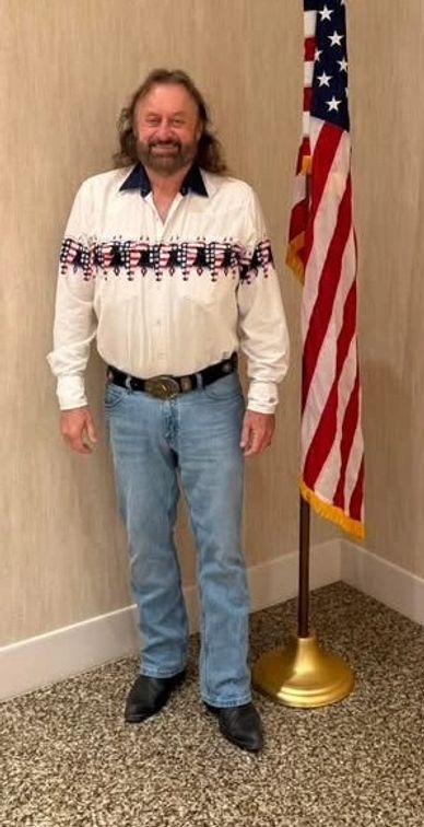 Man in patriotic shirt stands next to American flag indoors.