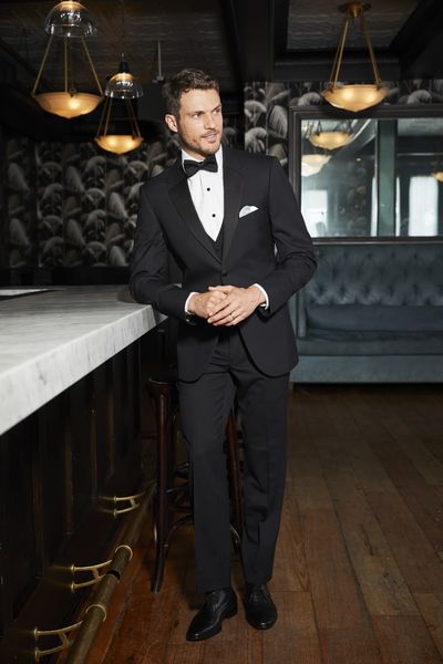 Man in a black tuxedo standing by a bar in an elegant room.