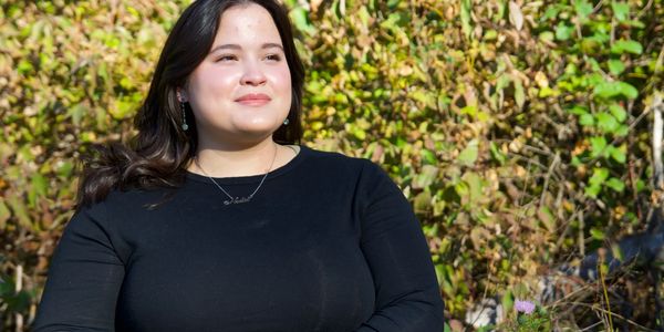 Woman in black top smiling outdoors with greenery background.