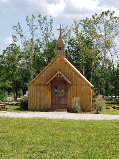 Small Rustic chapel with a cross on top in a green, wooded area.