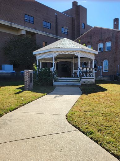 White gazebo with a weather vane in front of brick buildings under a clear blue sky.