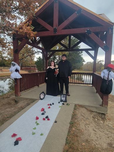 Couple dressed in black under a wooden gazebo with Halloween decorations.