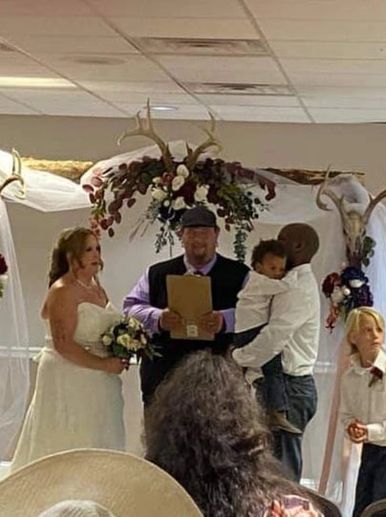 A wedding ceremony with bride, groom, child, and officiant under floral arch.