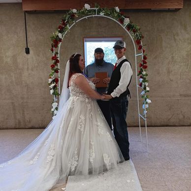 Bride and groom holding hands under a floral arch with officiant behind.
