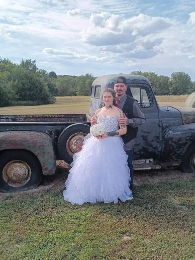 Couple posing in wedding attire by a rustic old truck in a countryside setting.