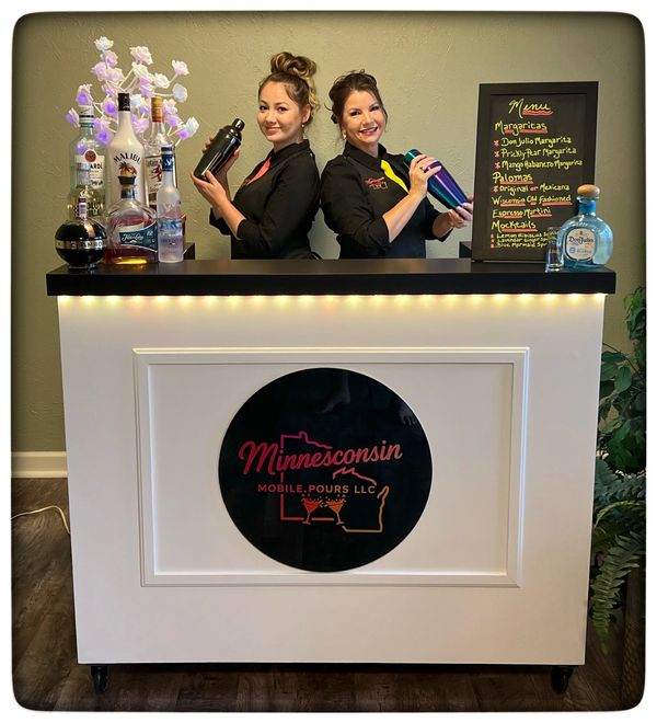 Two female bartenders stand behind a mobile bar with various liquor bottles and a cocktail menu.