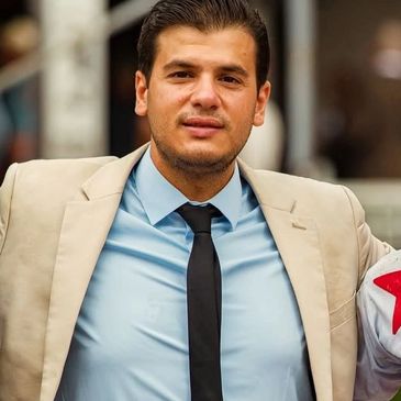 A confident man in a beige suit, blue shirt, and black tie.