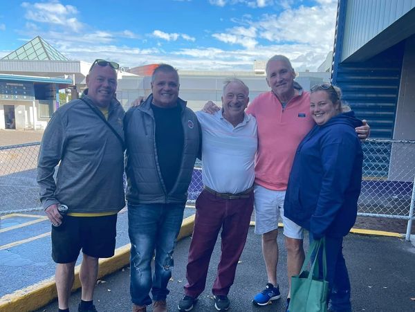 Five adults smiling outdoors on a sunny day, standing close together.