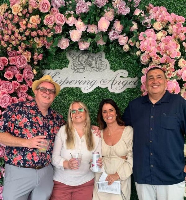 Four people smiling in front of a floral backdrop with 'Whispering Angels' sign.