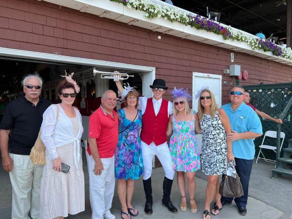Group of people posing outdoors in colorful summer attire with a man dressed in a horse racing jockey costume.