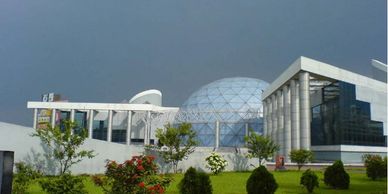 Modern building with a glass dome and greenery in front under a cloudy sky.
