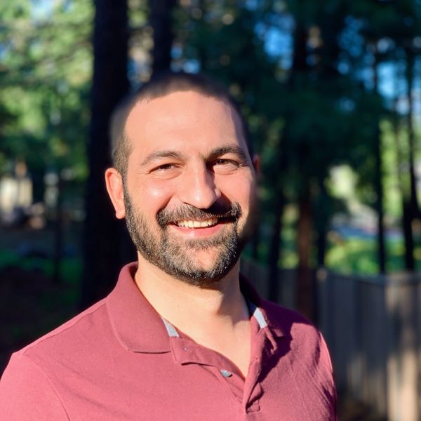 Smiling man in a maroon polo shirt outdoors with trees in the background.