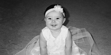 Smiling baby girl in a white dress and headband sitting on carpet.