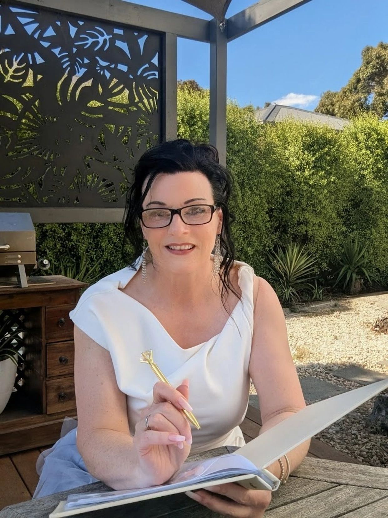 Woman in white dress smiling and holding a pen outdoors at a wooden table.