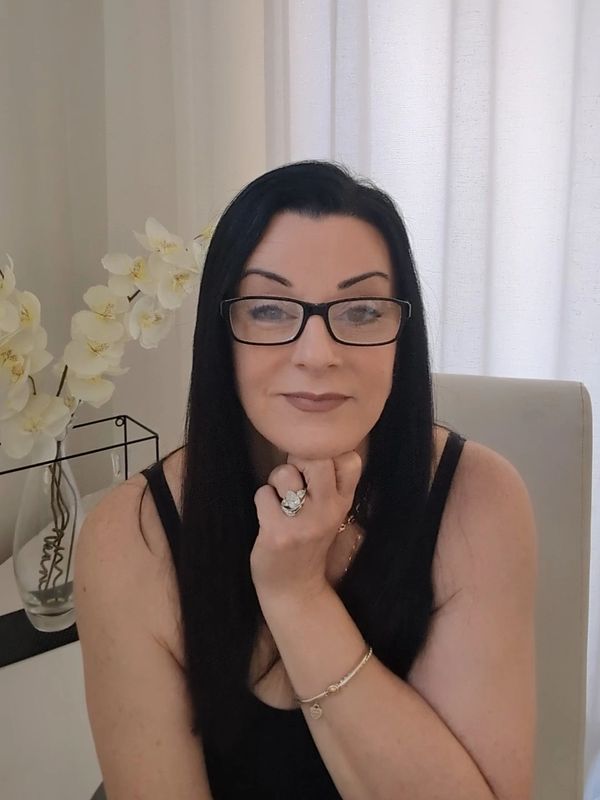 A woman with long black hair and glasses smiles gently indoors.