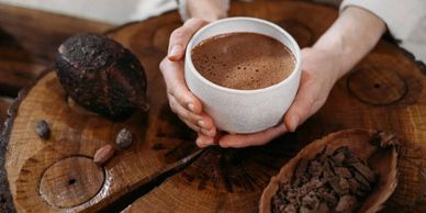 Hands holding a cup of hot chocolate on a wooden surface with cocoa beans.