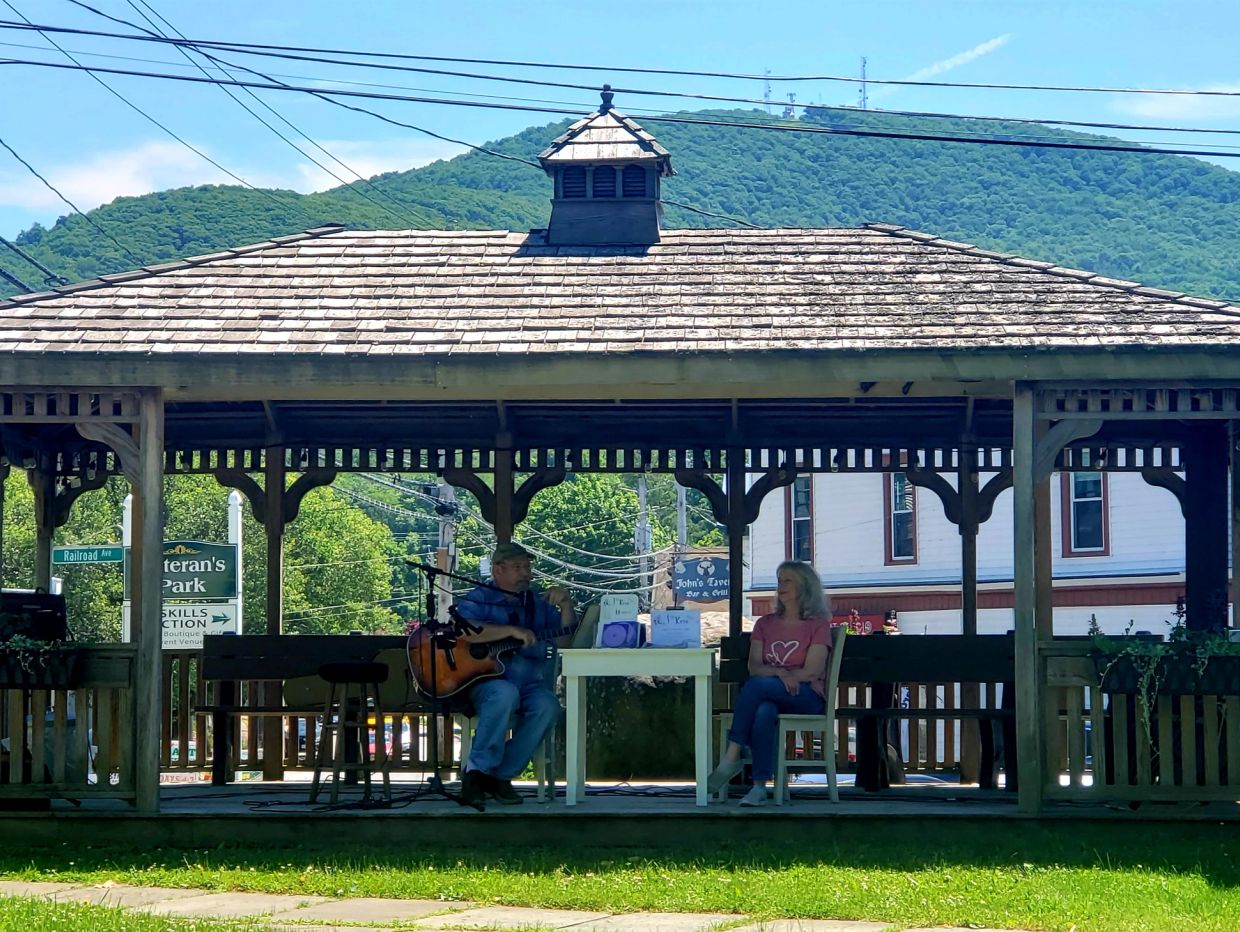 A man plays guitar and sings while a woman listens in a wooden gazebo.