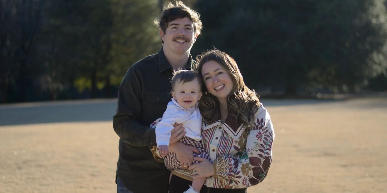 Smiling family of three posing outdoors on a sunny day.