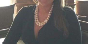 Woman wearing a black dress and pearl necklace sitting indoors.
