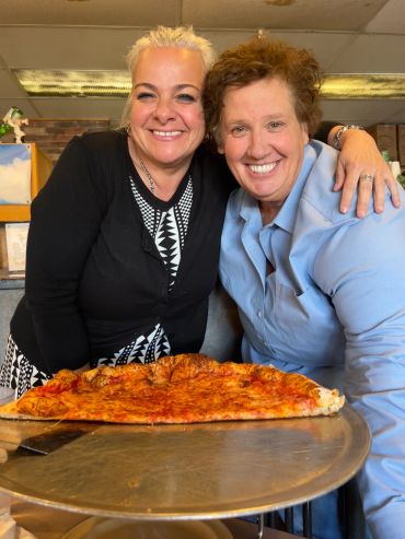 Two women smiling behind a large slice of pizza.