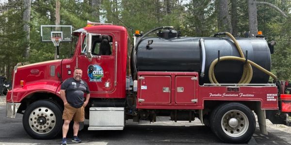 Man stands next to a red sanitation truck in a wooded area.