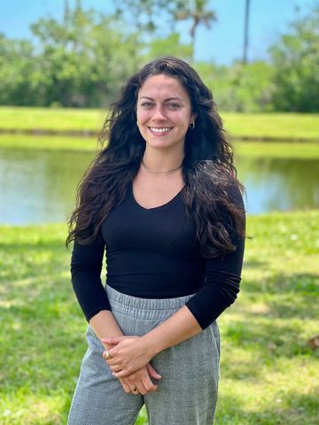 Woman with long dark hair standing outdoors by a pond, smiling at the camera.