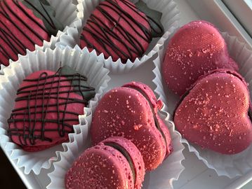 Assorted heart-shaped and round pink macarons with chocolate drizzle in a white box.