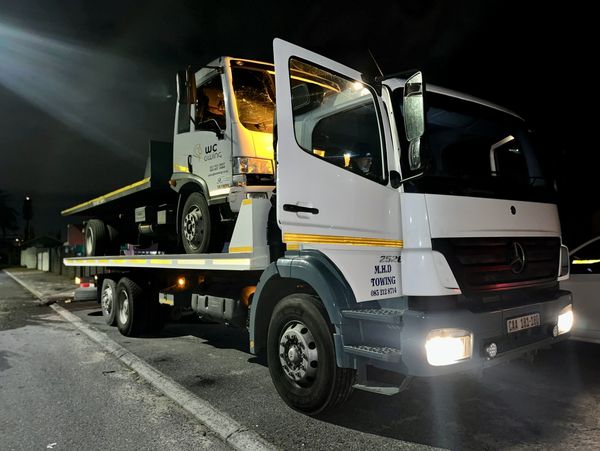 Tow truck carrying a damaged flatbed truck at night with headlights on.