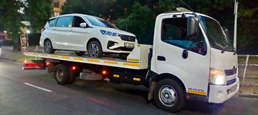 White MHD Transport truck towing a white SUV in the road Infront of some trees.