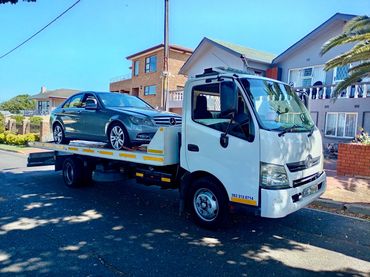 White MHD Transport tow truck parked in the road with a silver Mercedez on its deck.