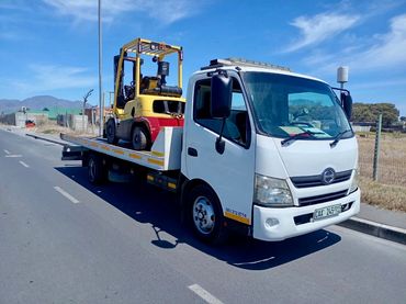 White MHD Transport truck loaded with a yellow forklift on its deck standing in the road.