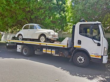 White MHD Transport truck loaded with a VW Beatle parked on its deck standing in the road.