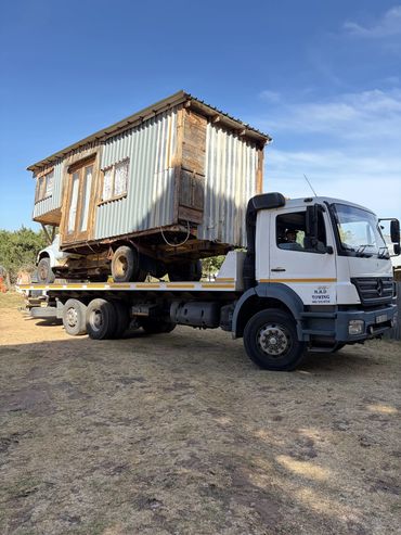 MHD Trasport flatbed tow truck transporting a large wooden cabin structure on rural terrain.
