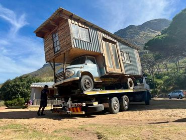 MHD Transport heavy-duty recovery truck carrying a wooden shack with mountains in the background.
