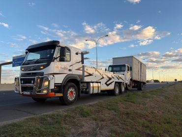 Tow truck hauling a white box truck on the roadside under a partly cloudy sky.