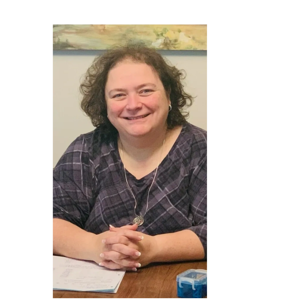 Angela is sitting at a desk and smiling with her hands folded in front of her. 
