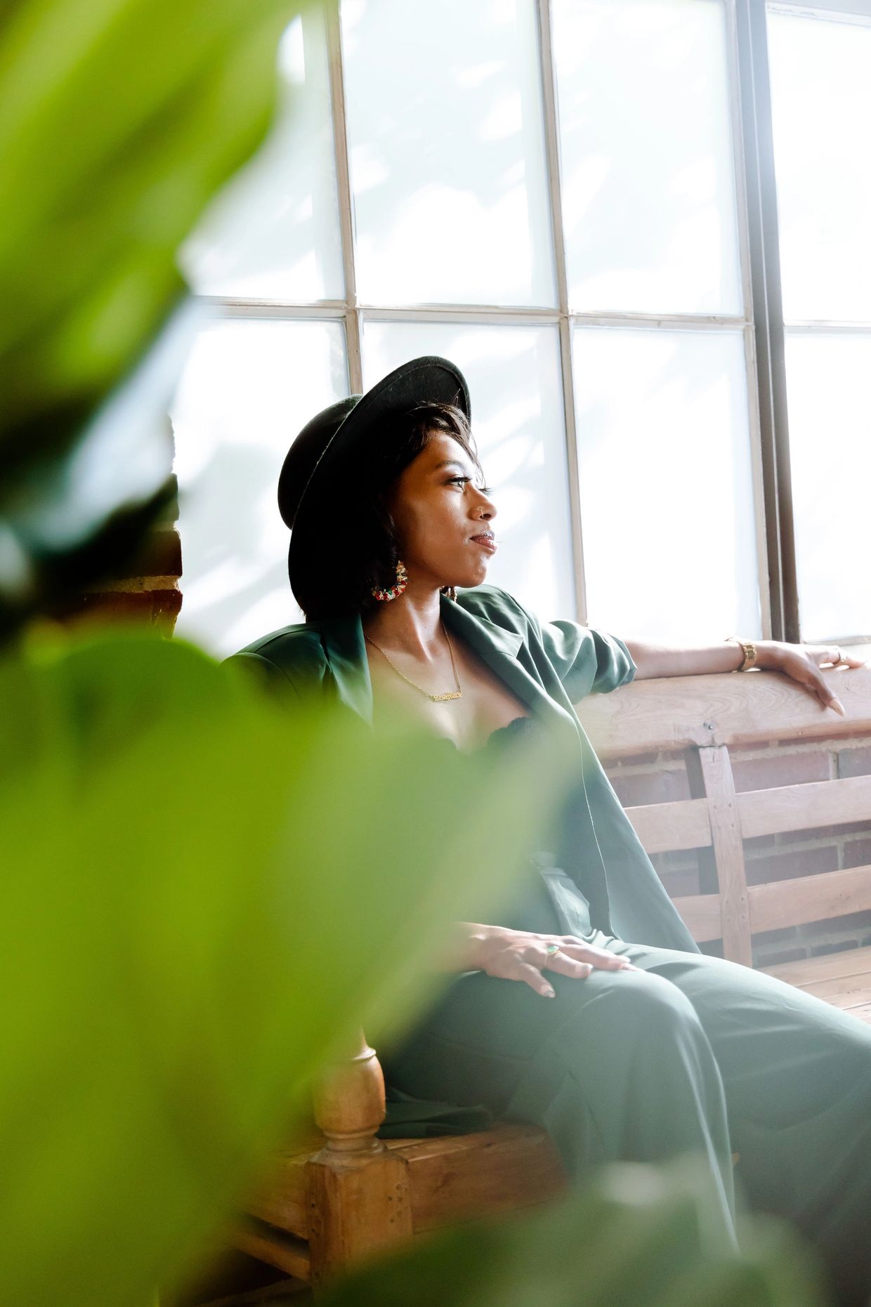 Woman in a stylish green suit and black hat relaxes by a bright window.