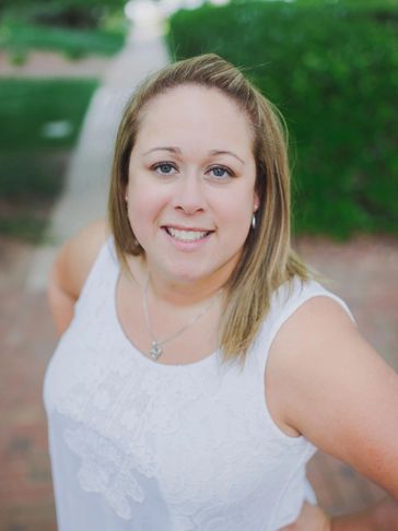 Smiling woman in white sleeveless top posing outdoors with greenery in the background.