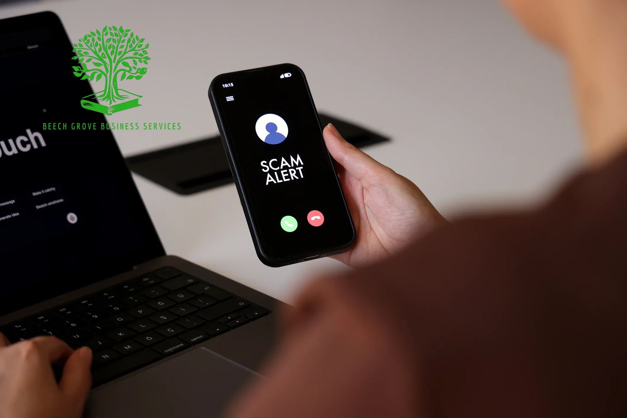 In an office environment, a worker in front of a laptop sees a message 'Scam Alert' on his mobile ph