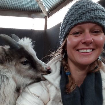 Woman smiling with a goat nuzzling her shoulder.