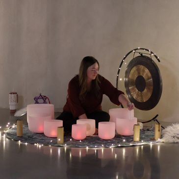 Woman playing crystal singing bowls in a serene setting with a gong and candles.