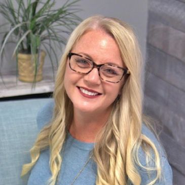 Smiling woman with long blonde hair and glasses sitting on a blue couch.