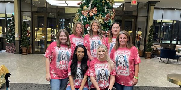 Seven women in matching festive shirts posing by a Christmas tree indoors.