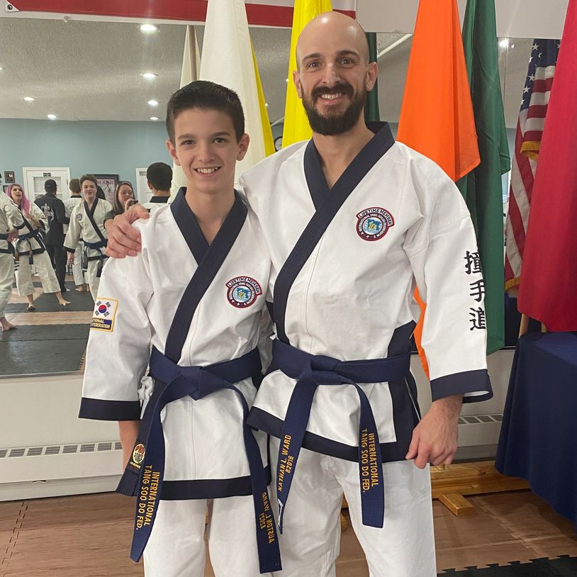 Two martial artists in white uniforms with blue belts pose together in a dojo.