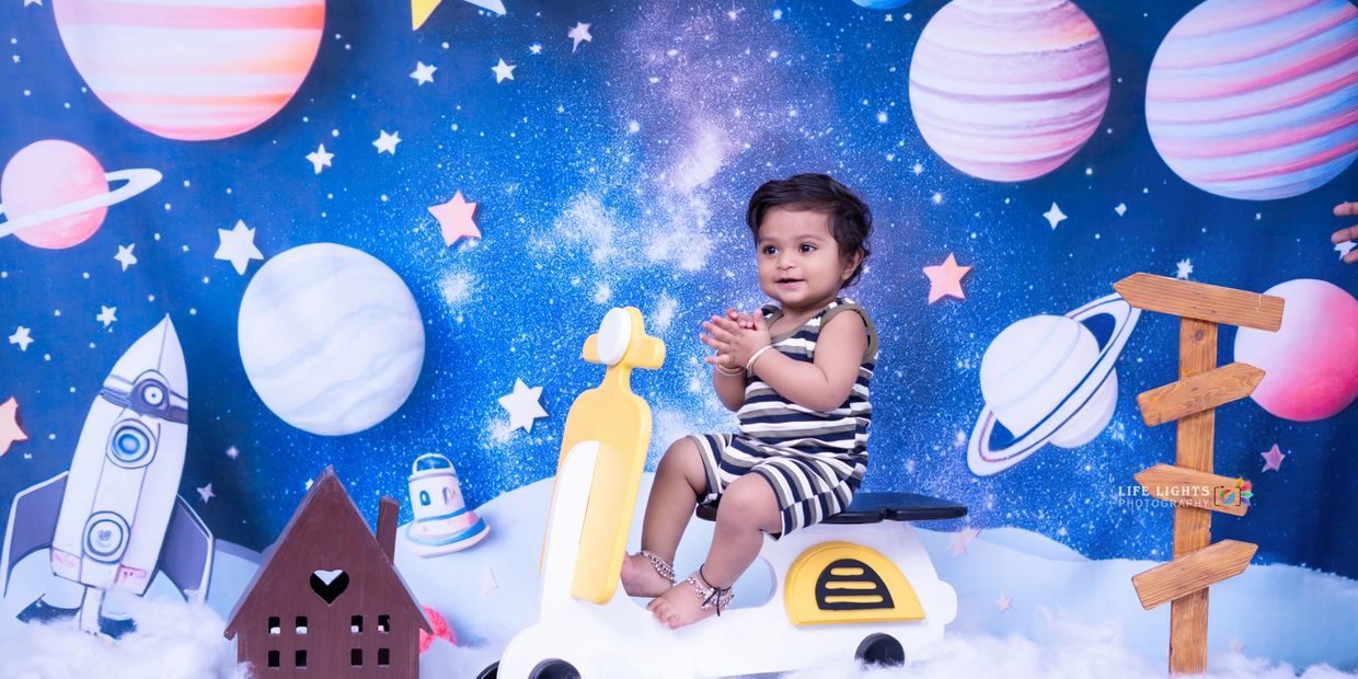 A happy baby sitting on a toy scooter with a space-themed backdrop.