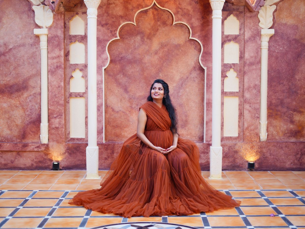 Pregnant woman in a flowing rust-colored dress sitting against ornate wall.