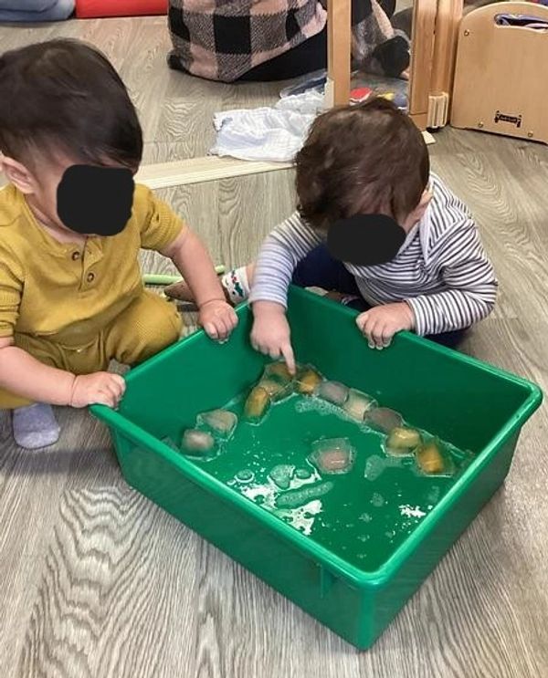 Two toddlers play with ice cubes in a green plastic tub on a wooden floor.