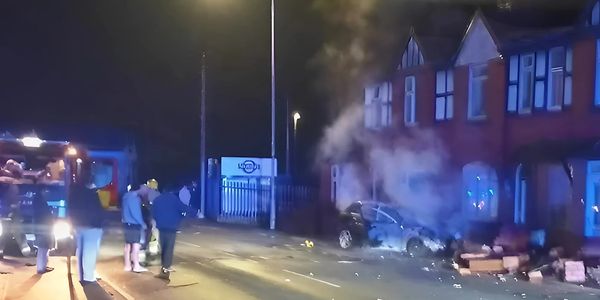 A car on fire near a row of houses at night with onlookers and emergency response.